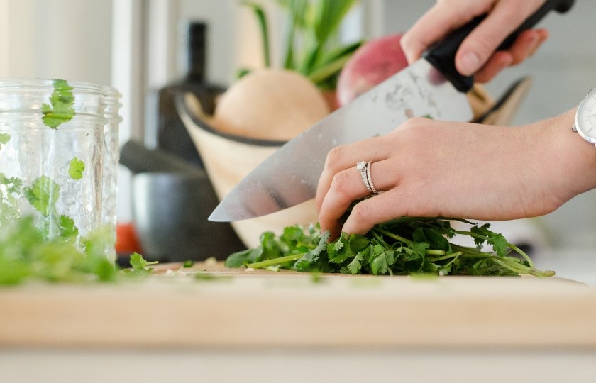 Close-up of a woman's hands chopping fresh herbs on a wooden cutting board, illustrating kitchen action verbs in Spanish.