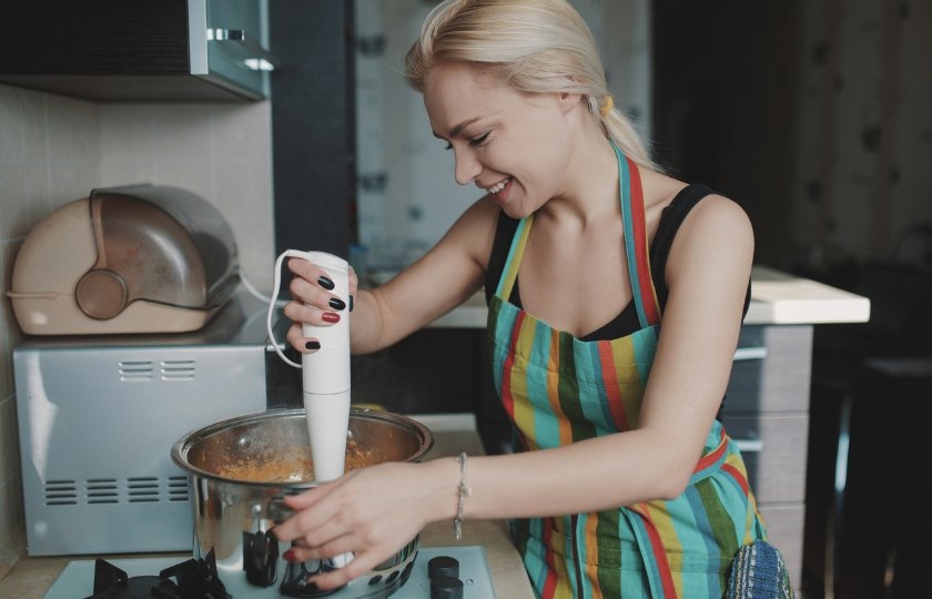 A woman smiling while using a hand blender, perfect for learning Spanish terms for everyday kitchen appliances.